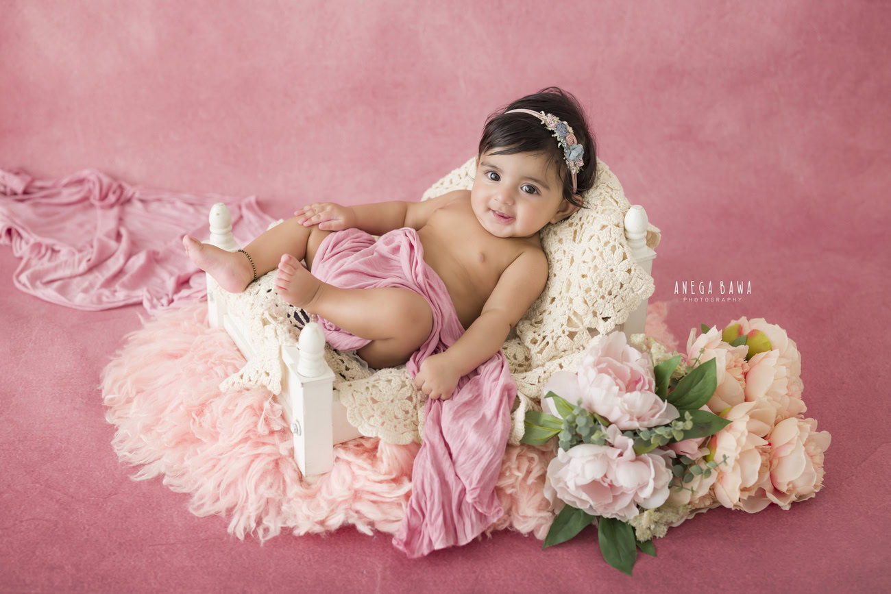 Baby girl photoshoot featuring a 3, 4, 5, or 6-month-old on a pink backdrop, wrapped in pink with delicate flowers. Contact Anega Bawa for sessions in Delhi, Gurgaon, or Noida.