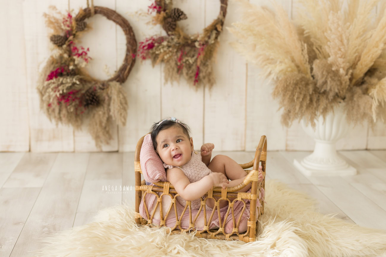 Capture the precious moments of your baby girl at 3, 4, 5, or 6 months in a charming setup with a wooden cot, beige rug, wooden frame, and brown shrubs against a beige backdrop. Book your session with Anega Bawa in Delhi, Gurgaon, or Noida.
