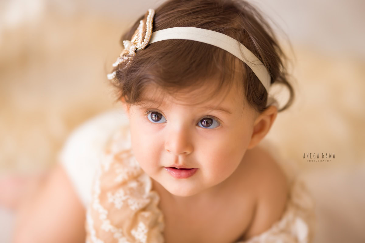 Captivating girl with beautiful eyes in front of a radiant golden backdrop, adorned with a golden headband, in her mesmerizing 10-11 months first birthday photoshoot in Delhi by Anega Bawa, Gurgaon, Noida