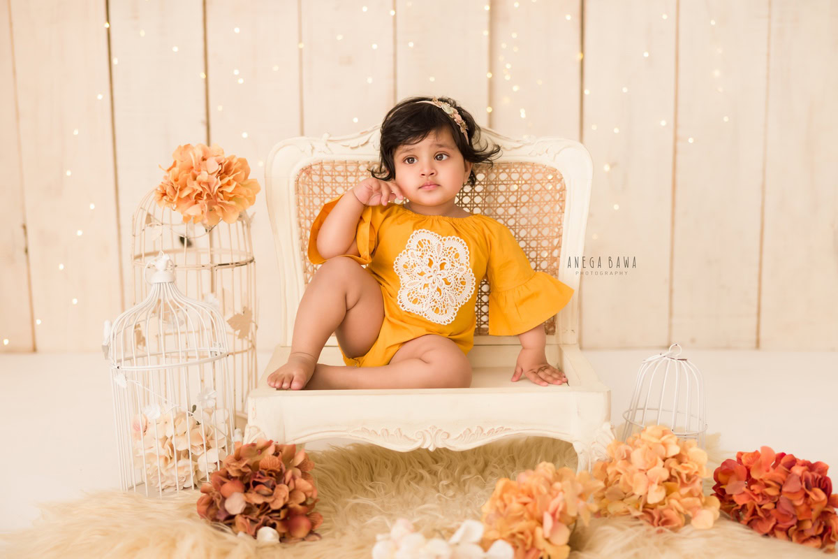 Stunning beige backdrop frames a 12-month-old girl sitting elegantly on a white wooden chair, donning a charming yellow dress amidst orange and brown flowers. This captivating image was skillfully captured by Anega Bawa, a talented sitter photographer in Gurgaon, Delhi.