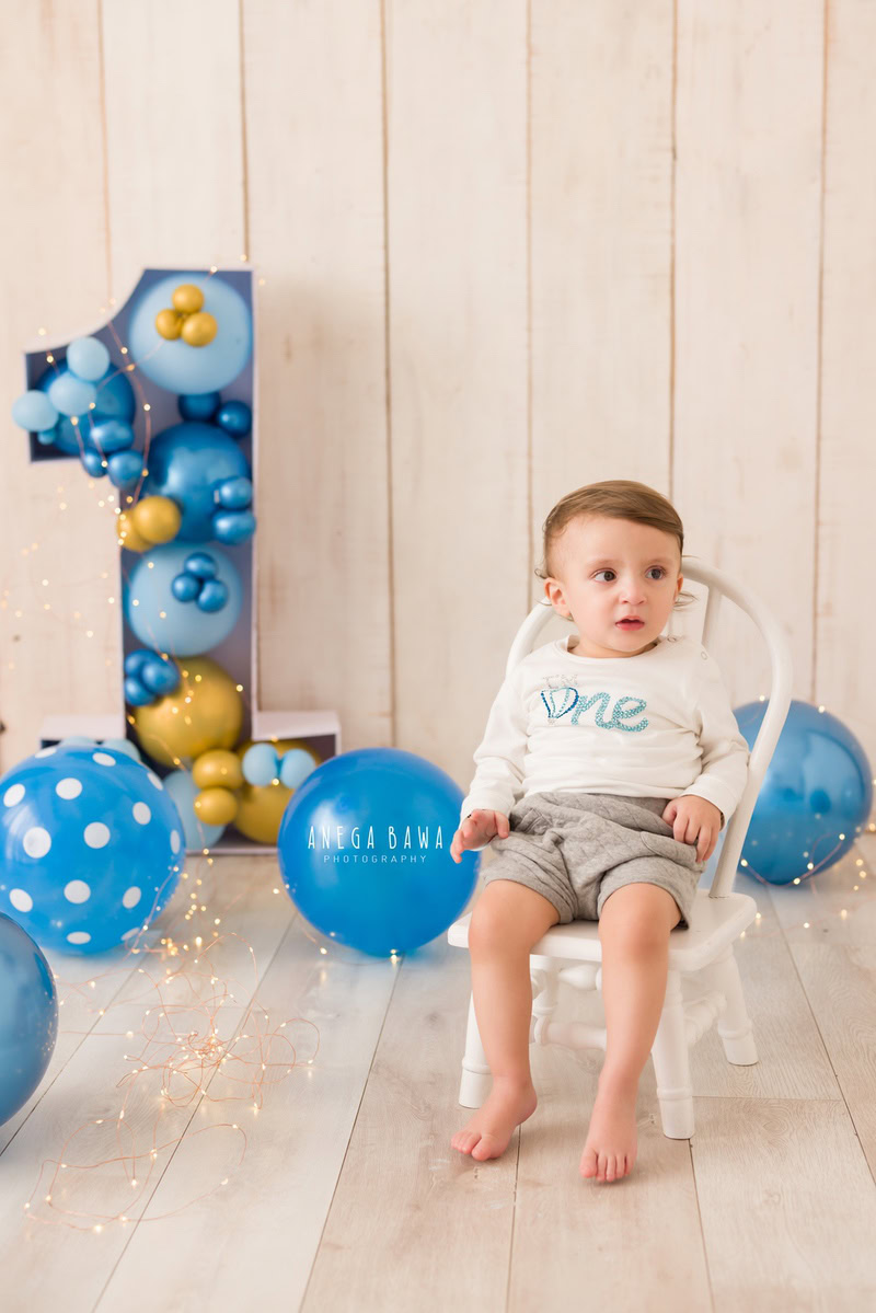 In this captivating scene from a baby pre-birthday photoshoot by Anega Bawa in Delhi, a handsome boy aged 10-11 months sits gracefully on a white chair against a soothing beige backdrop adorned with vibrant blue balloons. The combination of the serene backdrop and the playful pops of blue creates a charming ambiance, perfectly capturing the joy and innocence of childhood.