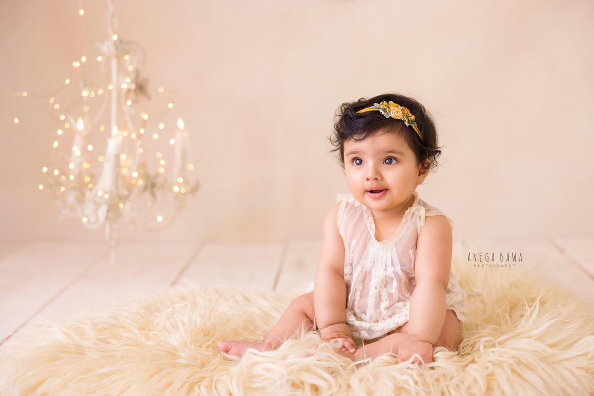 Girl seated on a beige rug against a charming beige backdrop, beside a candle stand emitting golden lights, captured during her memorable 10-11 months first birthday photoshoot in Delhi by Anega Bawa, Gurgaon, Noida