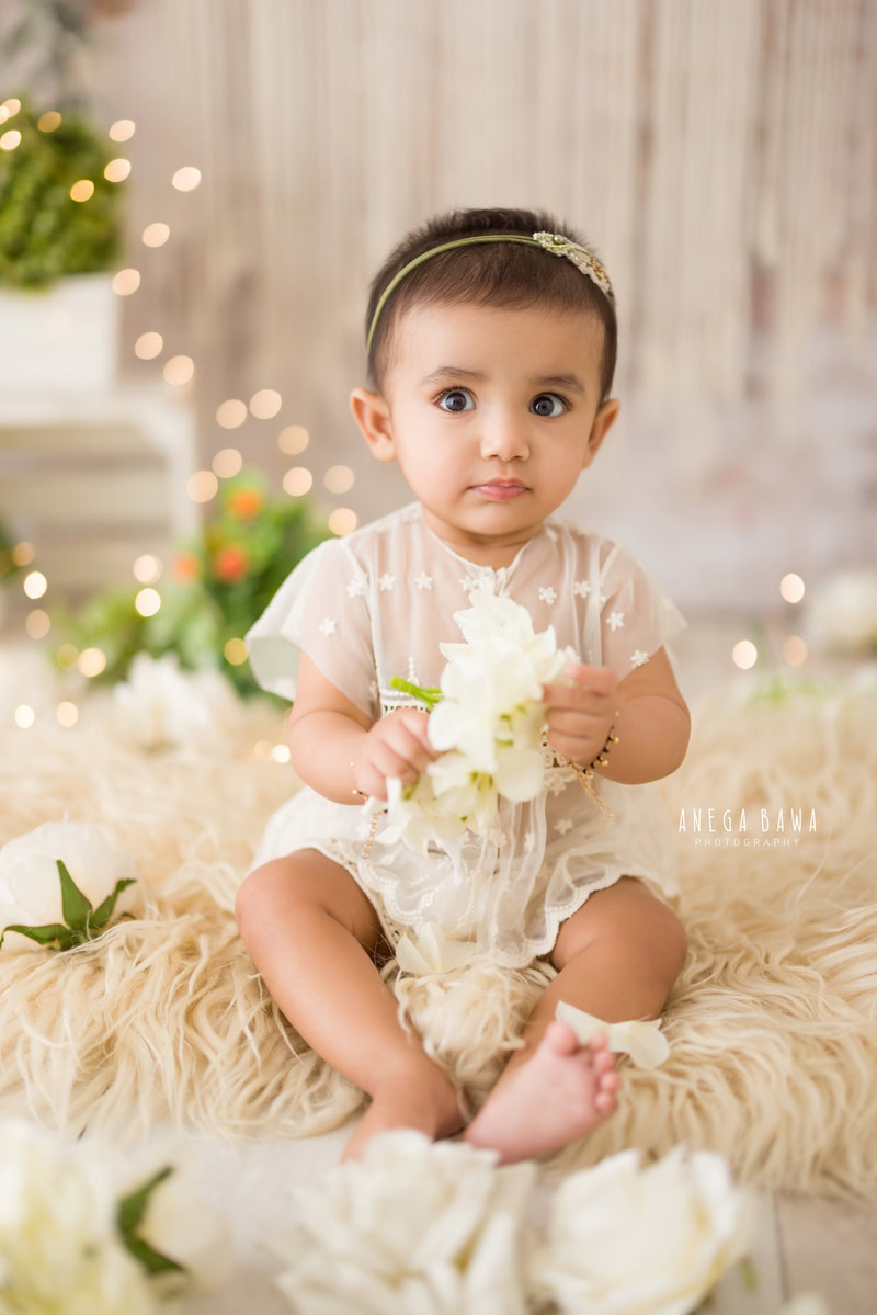 Girl sitting on a beige rug against a serene beige backdrop, surrounded by twinkling fairy lights, immortalizing her enchanting 10-11 months first birthday photoshoot in Delhi by Anega Bawa, Gurgaon, Noida