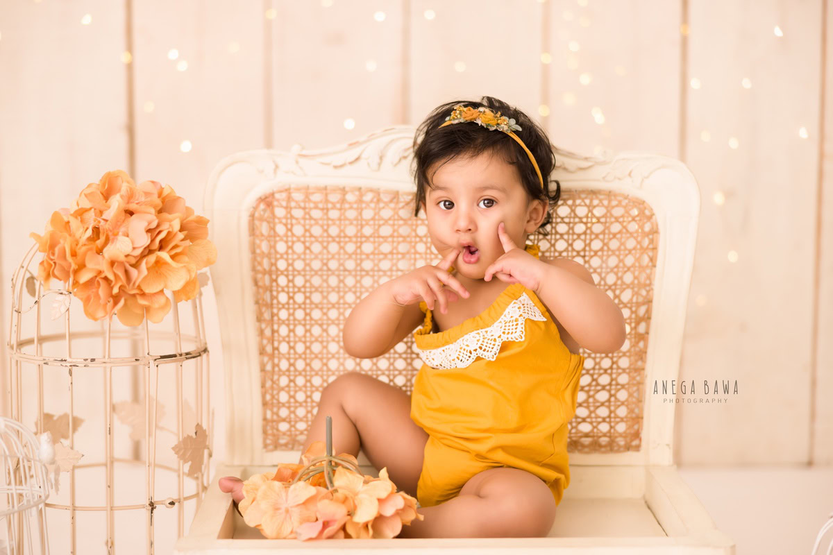 In front of a captivating beige backdrop illuminated with golden lights, a delightful 12-month-old girl sits gracefully on a wooden chair, surrounded by vibrant orange flowers. This enchanting moment was expertly captured by Anega Bawa, a skilled sitter photographer in Gurgaon, Delhi.