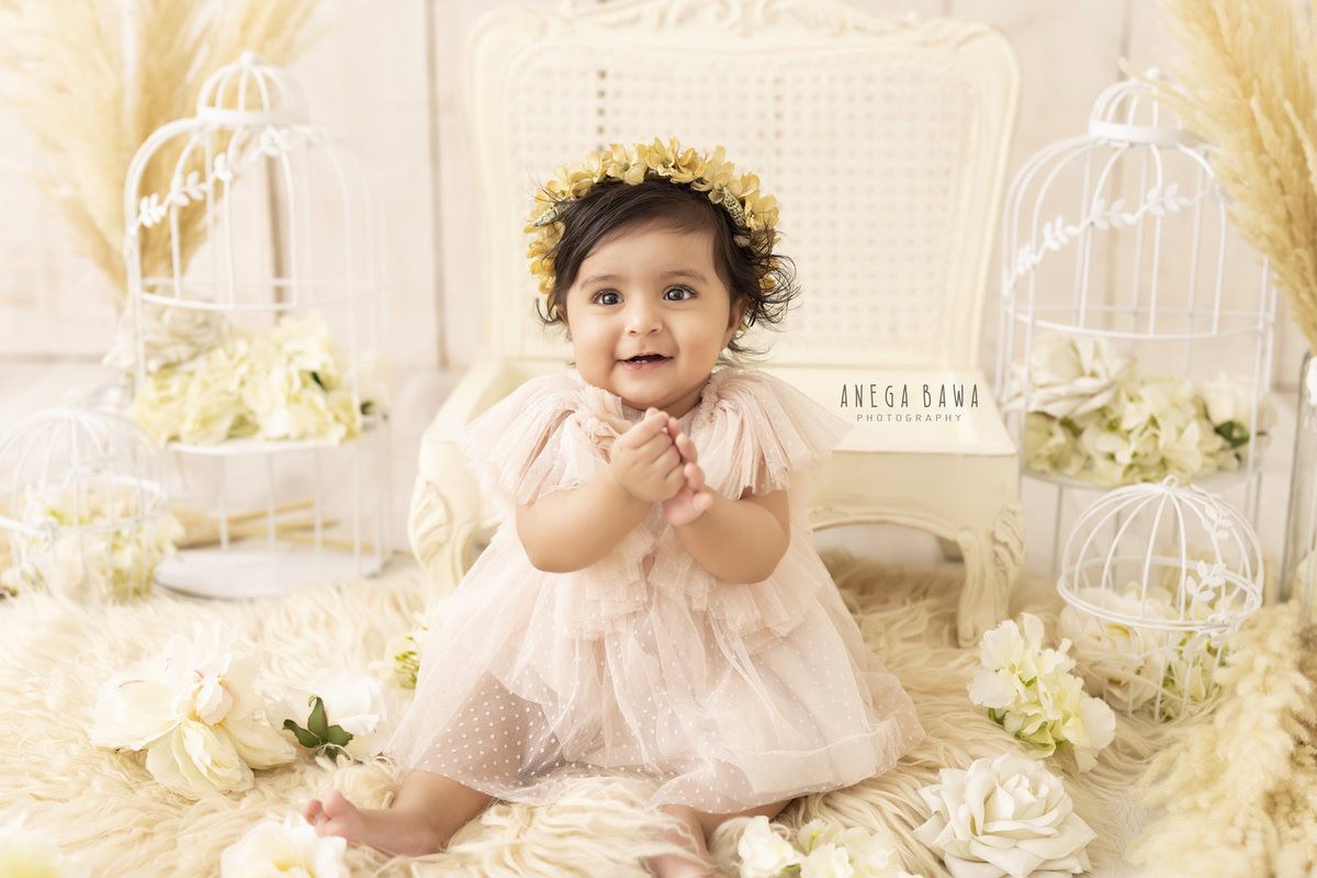 Serene beige backdrop adorned with white castles, showcasing a girl wearing a tiara band amidst delicate white flowers, capturing the magic of her 10-11 months first birthday photoshoot in Delhi by Anega Bawa, Gurgaon, Noida