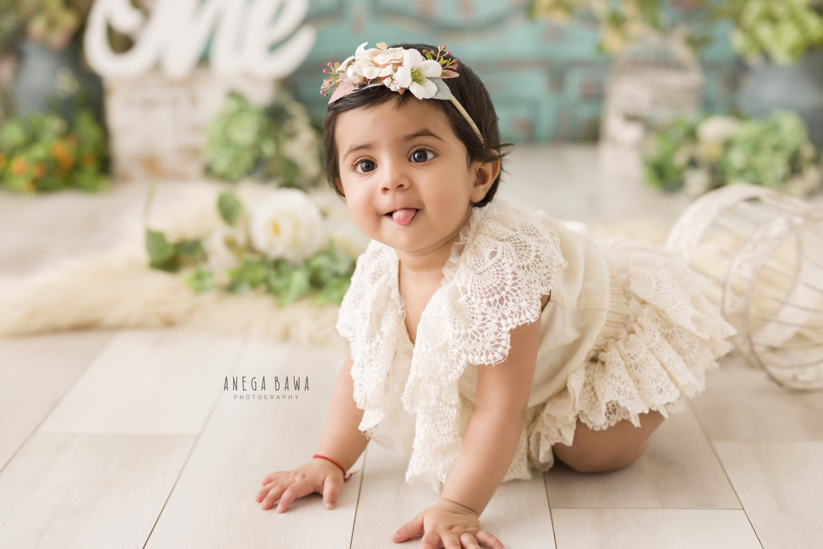 A serene beige backdrop embellished with delicate white flowers, showcasing a one-year-old girl in a charming net dress surrounded by enchanting castles, captured during her 10-11 months first birthday photoshoot in Delhi by Anega Bawa, Gurgaon, Noida