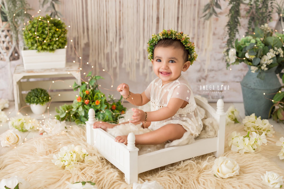 A cozy beige rug illuminated by twinkling fairy lights, featuring a girl sitting in a white cot adorned with a tiara band, surrounded by white pots, capturing the essence of her 10-11 months first birthday photoshoot in Delhi by Anega Bawa, Gurgaon, Noida