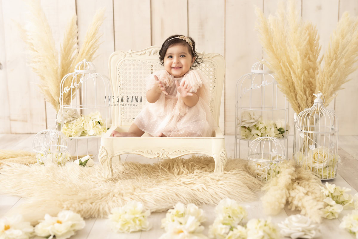 Behold the enchanting sight of a smiling 1-year-old girl seated on a white wooden chair, surrounded by a beige rug and white castles. With brown shrubs and white flowers adorning the floor, this picturesque moment was expertly captured during a toddler photoshoot in Delhi, Gurgaon, and Noida by the talented photographer, Anega Bawa.