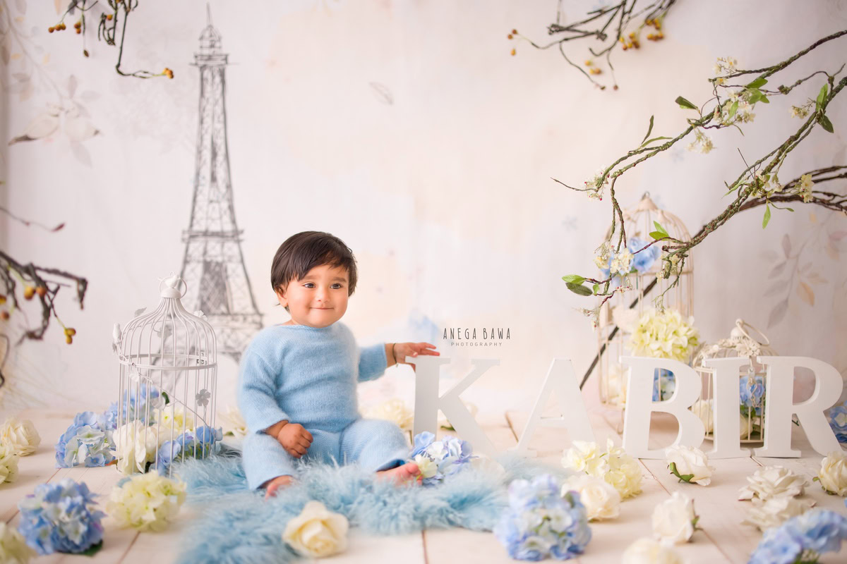 Celebrate your little boy's 10-11 months milestone with a charming sitter photoshoot by Anega Bawa Photography in Delhi and Gurgaon. Dressed in a blue baby suit, seated on a soft blue rug against an Eiffel Tower wallpaper backdrop, each frame captures his adorable presence alongside a personalized name frame on the floor, creating cherished memories to last a lifetime.