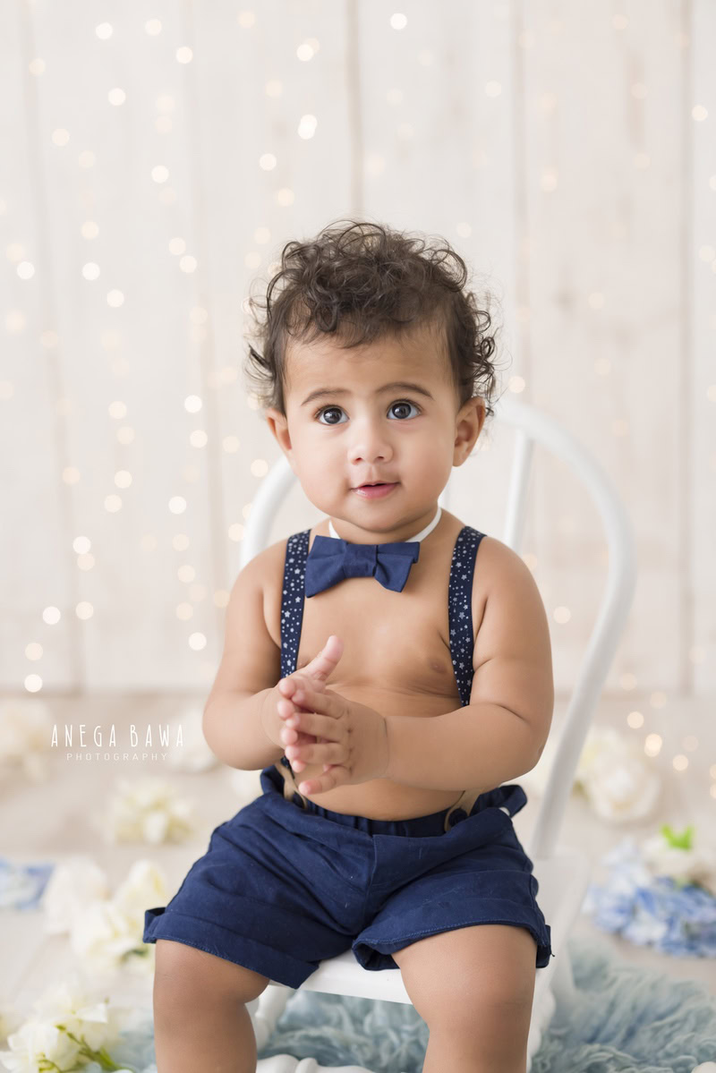 One-year-old boy sitting on a white chair atop a blue rug against a serene beige backdrop, in his memorable first birthday photoshoot in Delhi by Anega Bawa, Gurgaon, Noida