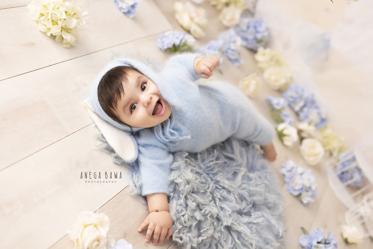 Charming boy dressed in a blue babysuit and matching cap, seated on a blue rug against a serene beige backdrop, captured during his delightful 8-9 months first birthday photoshoot in Delhi by Anega Bawa, Gurgaon, Noida
