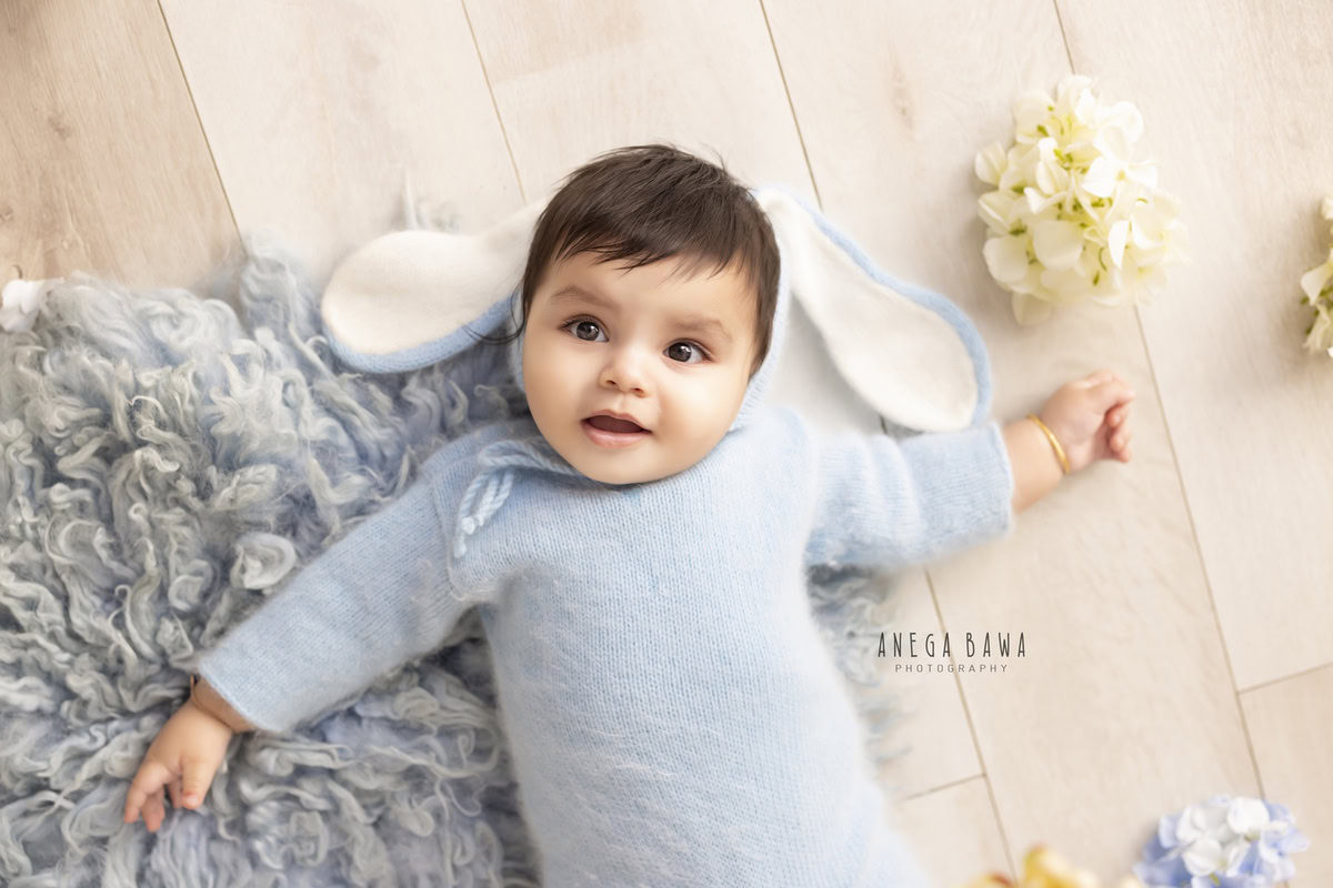 Immerse yourself in the delightful innocence of a boy clad in a blue babysuit, seated on a blue rug against a serene beige backdrop. With delicate flowers adorning the floor, this captivating scene was skillfully captured during a toddler photoshoot in Delhi, Gurgaon, and Noida by the gifted photographer, Anega Bawa.