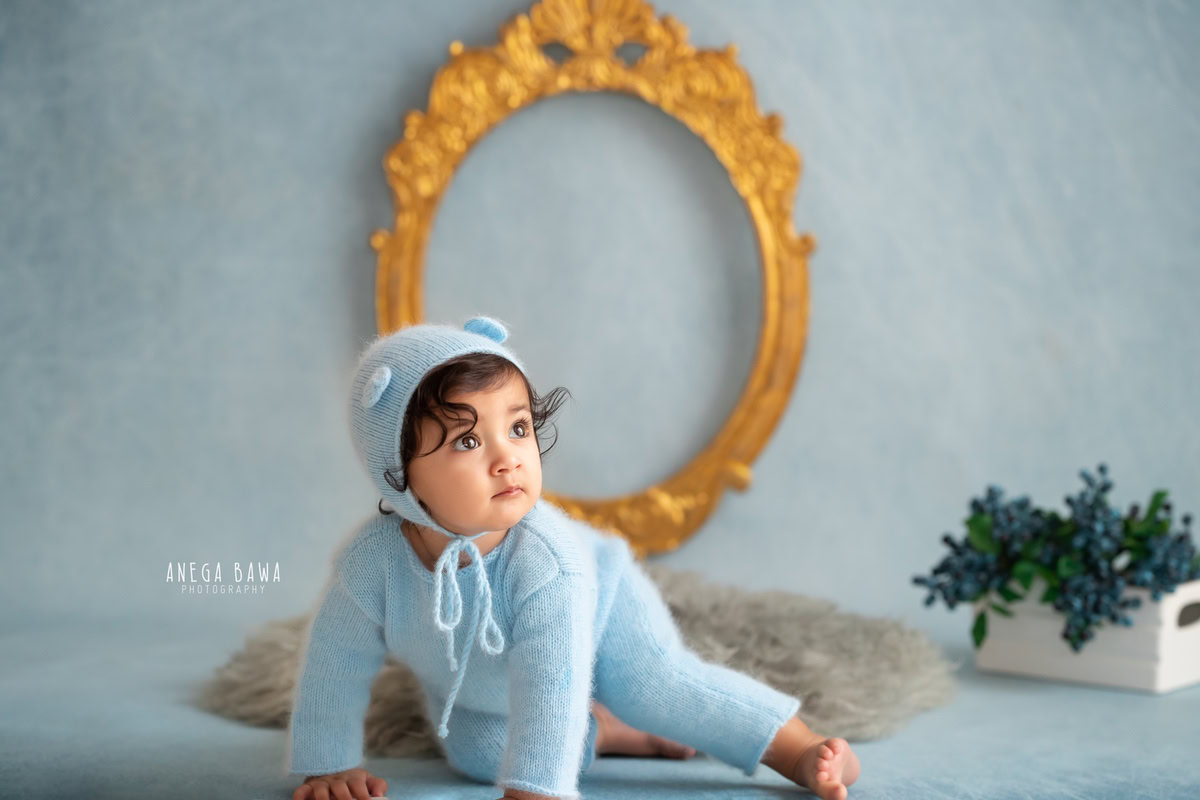 Embrace the charm of a little boy clad in a blue baby suit, sporting a cute cap, set against a serene blue backdrop. With a golden frame adorning the wall and a soft grey rug beneath, this scene captures the innocence of a 8-9 months old during a sitter photoshoot in Delhi and Gurgaon.