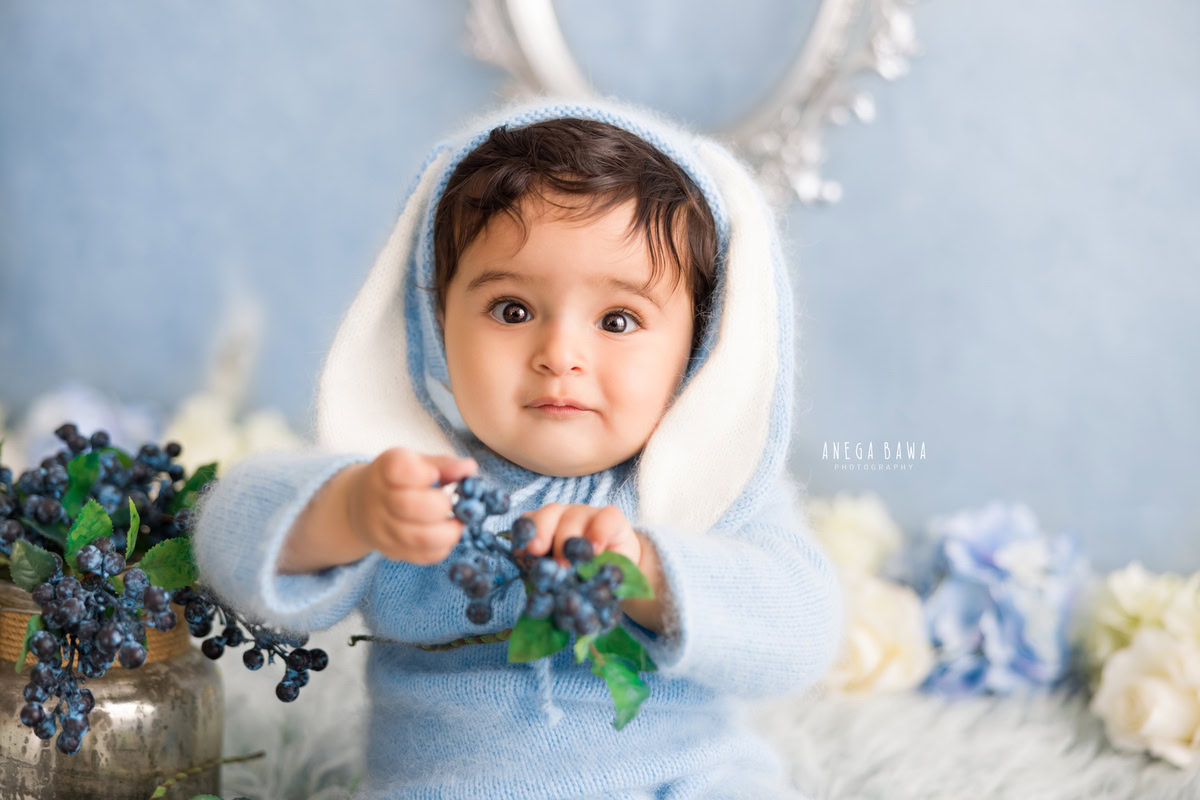 Delight in the adorable sight of a young boy dressed in a charming blue baby suit, accentuated by a cute cap. Against a backdrop reminiscent of blueberries, complemented by a soft grey rug, this scene encapsulates the innocence of a 8-9 months old during a sitter photoshoot in Delhi and Gurgaon.