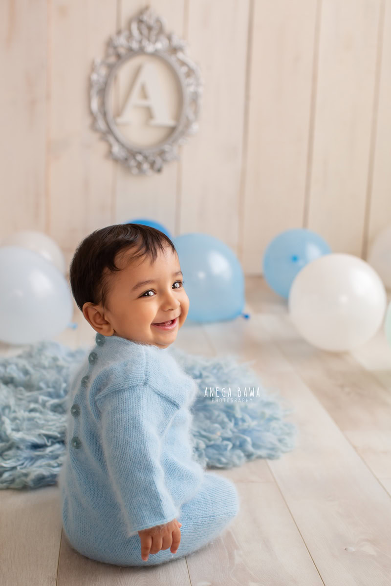 Dressed in a charming blue babysuit, a 8-9 months old boy poses next to a silver alphabet frame, surrounded by cheerful balloons, on a soft blue rug against a serene beige backdrop. This captivating moment was skillfully captured by Anega Bawa, a talented sitter photographer in Gurgaon, Delhi.