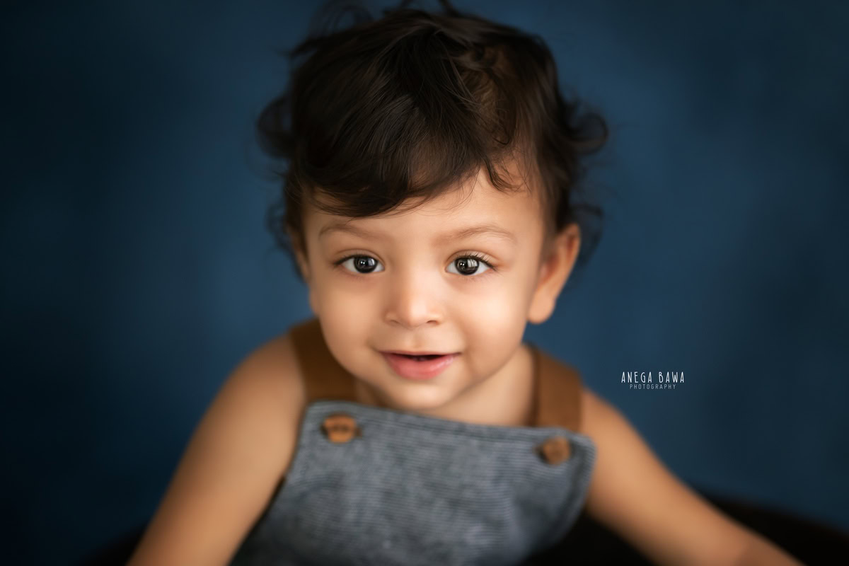 In adorable blue dungarees, a 8-9 months old boy stands against a vibrant blue backdrop, captured skillfully by Anega Bawa, a talented sitter photographer in Gurgaon, Delhi.
