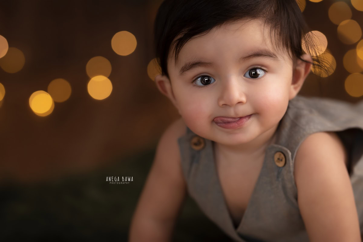 Bathed in the warm glow of golden lights, an 8-9 months old boy poses against a rustic brown backdrop, skillfully photographed by Anega Bawa, a renowned sitter photographer based in Gurgaon, Delhi.