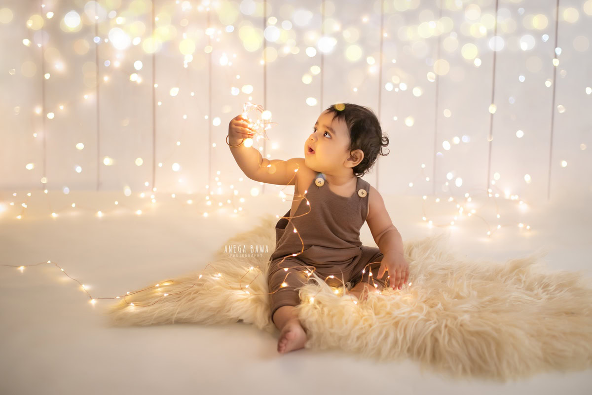 Create timeless memories of your 8-9-month-old boy with a sitter photoshoot by Anega Bawa Photography in Delhi and Gurgaon. Adorned in a charming brown dungaree, he sits on a cozy beige rug surrounded by enchanting fairy lights, set against a warm beige backdrop, capturing the innocence and joy of this special moment.