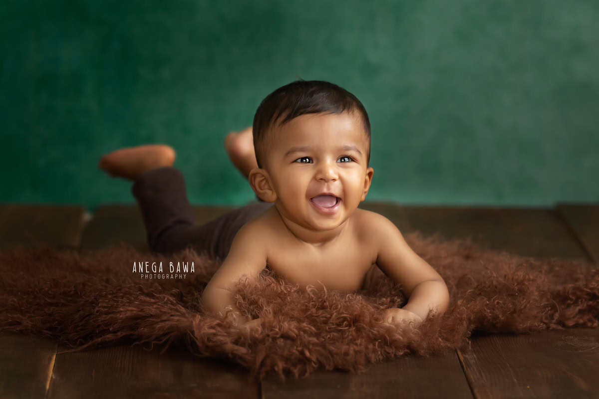 In a charming scene captured by Gurgaon's talented sitter photographer, Anega Bawa, a delightful 8-9 months old boy sits on a brown rug against a vibrant green backdrop in Delhi.
