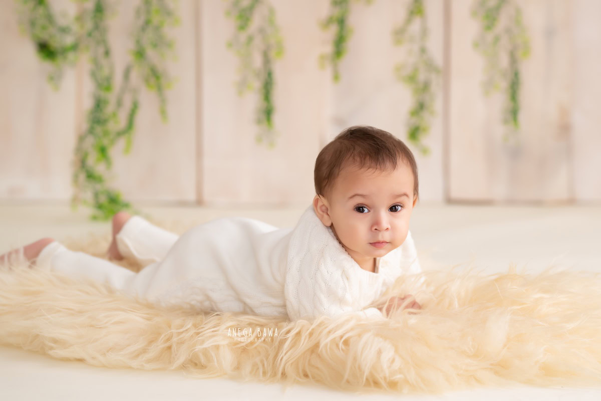In a captivating photo taken by the skilled sitter photographer Anega Bawa in Delhi, a little boy, aged 8-9 months, crawls on a soft beige rug, with a backdrop of beige and delicate leaves hanging on the wall, creating a serene atmosphere.