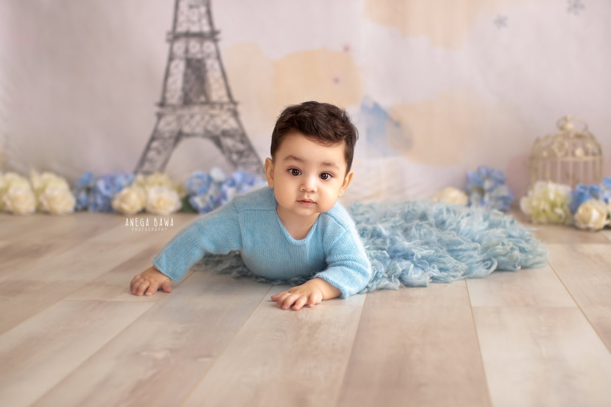 Capture the delightful moments of your 8-9-month-old boy's exploration with a charming sitter photoshoot by Anega Bawa Photography in Delhi and Gurgaon. Against a picturesque backdrop featuring the Eiffel Tower, dressed in a cozy blue babysuit, watch as he crawls on a soft blue rug, surrounded by golden castles, creating memories that will last a lifetime.