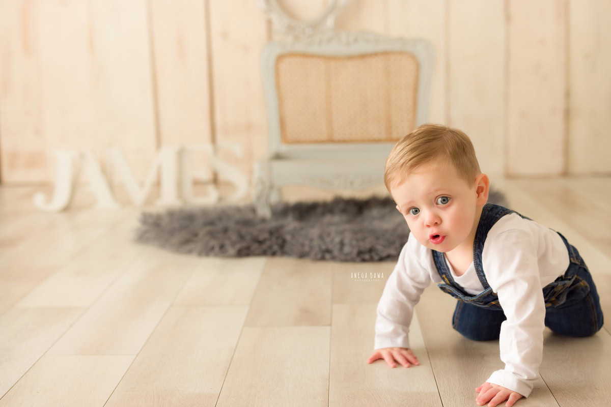 In this delightful capture from a baby pre-birthday photoshoot by Anega Bawa in Delhi, a sweet 8-month-old boy explores his surroundings with curiosity, crawling near a stylish grey wooden chair. The scene is set against a timeless beige backdrop, with a charming name frame resting on the floor nearby. This heartwarming image beautifully encapsulates the innocence and wonder of early childhood moments.