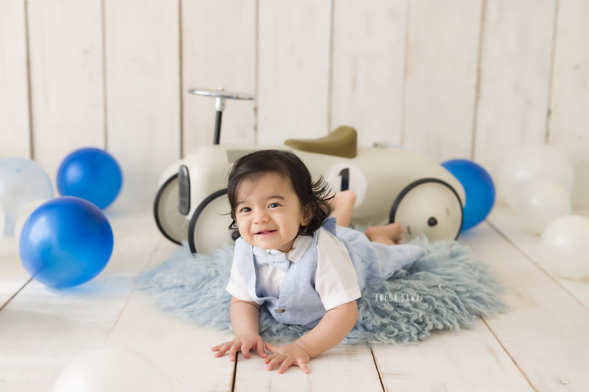 Delight in your little one's adventures with Anega Bawa Photography's sitter photography in Delhi and Gurgaon. Watch as he explores the world on a blue rug, with a toy scooter by his side, against a soothing beige backdrop adorned with blue balloons. These precious moments, captured at eight months old, are bound to become cherished memories.