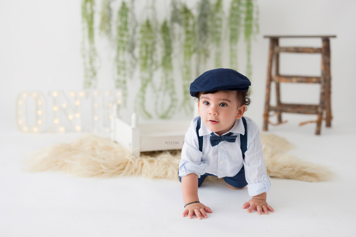 Adorable boy joyfully crawling on a beige rug with a leafy fringe on the wall, against a pristine white backdrop, with a charming name frame on the floor, celebrating his delightful 8 months first birthday photoshoot in Delhi by Anega Bawa, Gurgaon, Noida