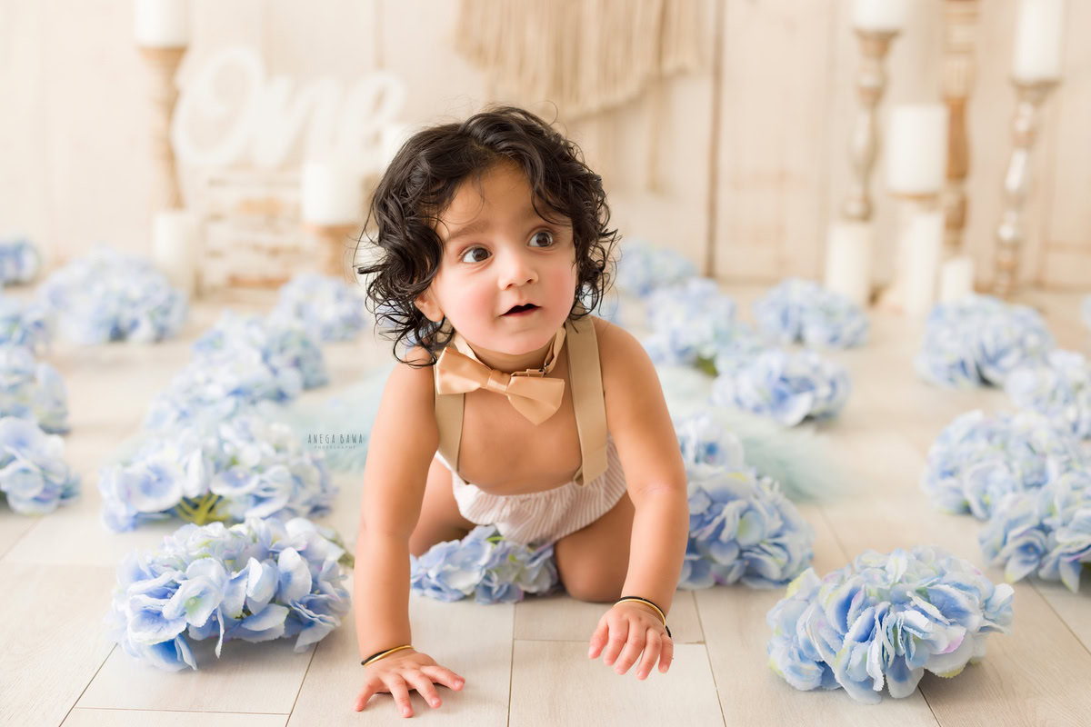 Captured during a baby pre-birthday photoshoot by Anega Bawa in Delhi, this delightful image features an 8-month-old boy joyfully crawling amidst vibrant blue flowers, adding a pop of color to the scene. Against a calming beige backdrop, the warm glow of a candle stand adds a touch of coziness to the atmosphere, creating a serene and enchanting ambiance.