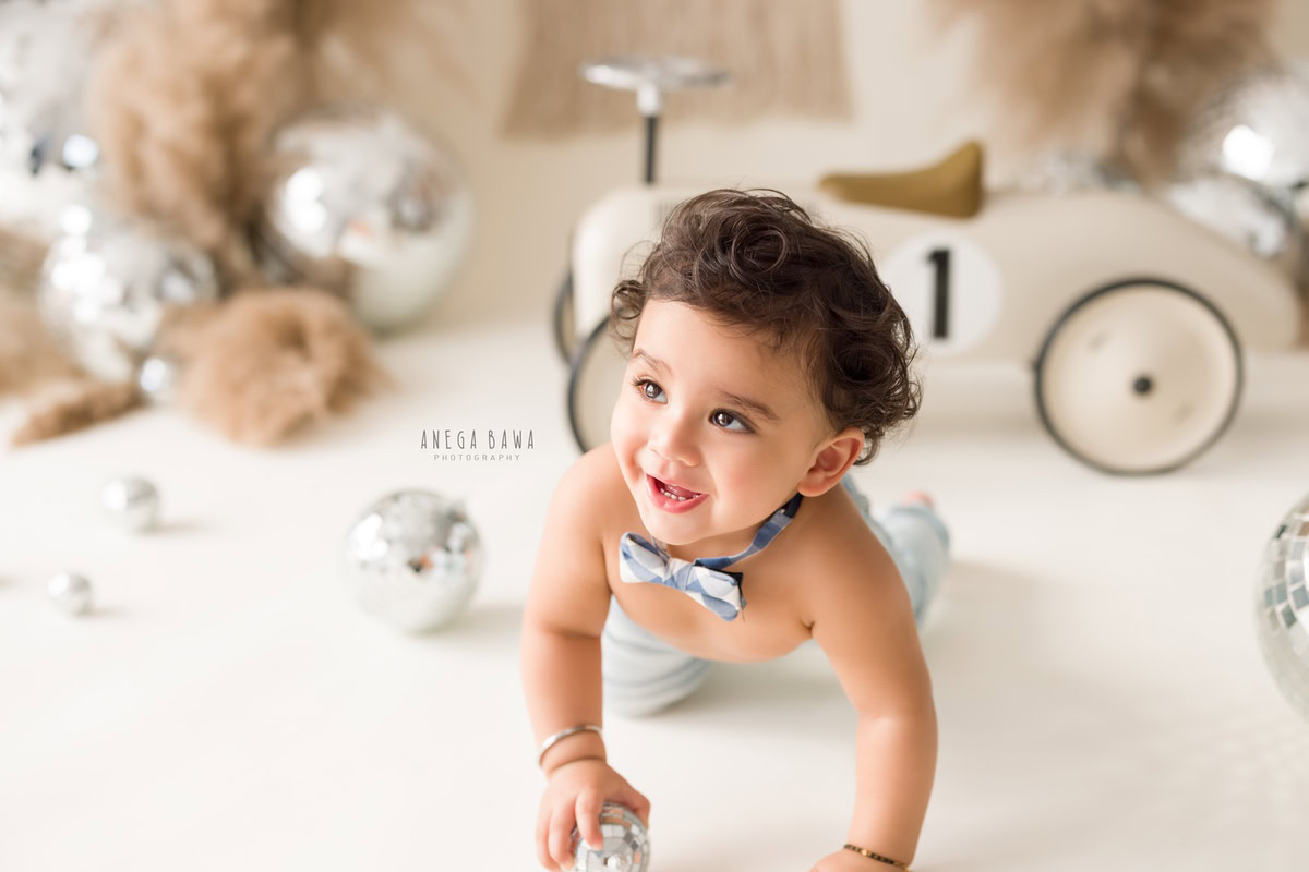 Amidst a captivating scene captured by esteemed sitter photographer Anega Bawa in Delhi, an adorable 8-month-old boy joyfully crawls on the floor, accompanied by a toy scooter and disco ball, amidst brown bushes, all set against a pristine white backdrop.