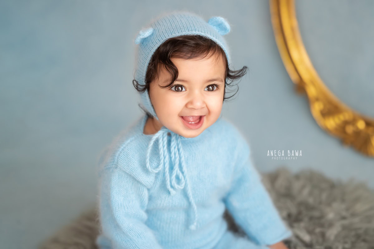 Captured during a sitter photo shoot in Delhi and Gurgaon by the talented Anega Bawa, this endearing 9-month-old boy, adorned in a cute cap, poses against a soothing blue backdrop. His charming innocence shines through as he sits on a soft grey rug, creating a timeless moment to cherish.