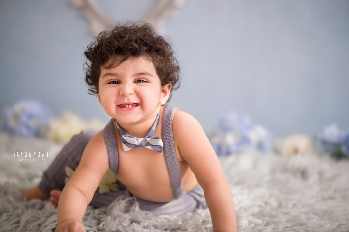 In this delightful sitter photo shoot captured by Anega Bawa in Delhi, Gurgaon, a charming 9-month-old boy steals the show in his grey dungaree against a captivating blue backdrop. The scene is adorned with a cozy grey rug and vibrant flowers, adding to the whimsical charm of the setting.