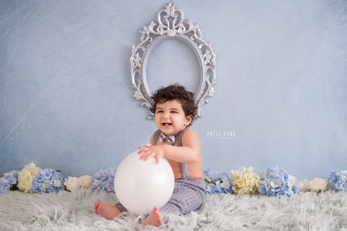 In this heartwarming sitter photo shoot by Anega Bawa in Delhi, Gurgaon, a joyful 9-month-old boy is captured holding balloons while seated on a soft grey rug against a serene blue backdrop. The scene is beautifully complemented by a grey frame on the wall and a cute bow, adding to the adorable charm of the moment.