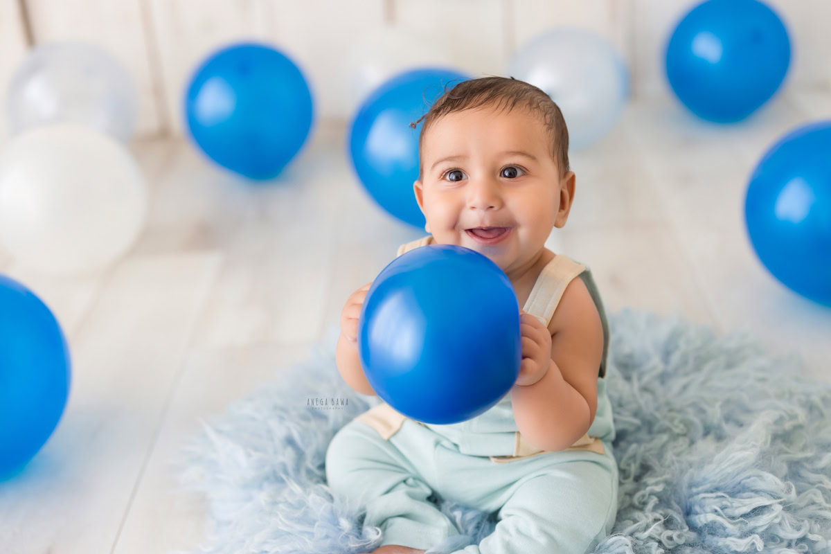Experience the joyous charm of a 9-month-old boy, cradling a vibrant balloon while seated on a soft blue rug against a pristine white backdrop. This heartwarming moment was beautifully captured during a toddler photoshoot in Delhi, Gurgaon, and Noida by the talented photographer, Anega Bawa.