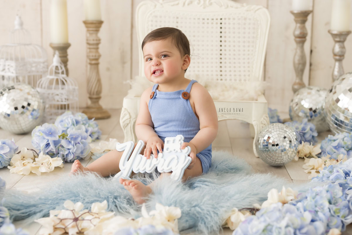 In this captivating shot from a baby pre-birthday photoshoot by Anega Bawa in Delhi, a 9-month-old boy holds a charming name frame, adding a personal touch to the scene. Set against a soothing beige backdrop, the scene is illuminated by the warm glow of a candle stand and disco balls, while a vibrant blue rug adds a delightful pop of color, creating a magical and festive ambiance.