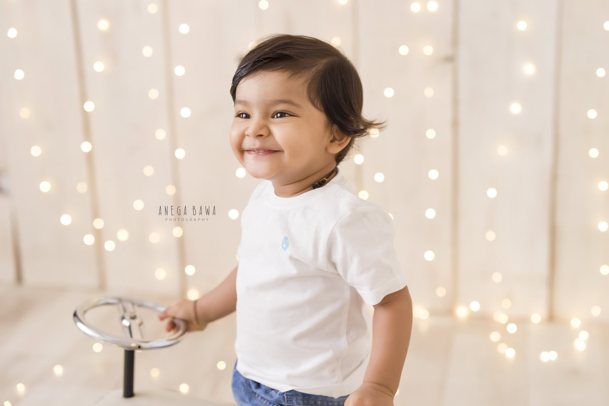 In this enchanting capture by Anega Bawa for a baby pre-birthday photoshoot in Delhi, a 9-month-old boy holds the wheel of a toy car, his eyes sparkling with curiosity and joy. Set against a serene beige backdrop, the scene is adorned with twinkling fairy lights, casting a soft glow and adding a touch of magic to the moment.