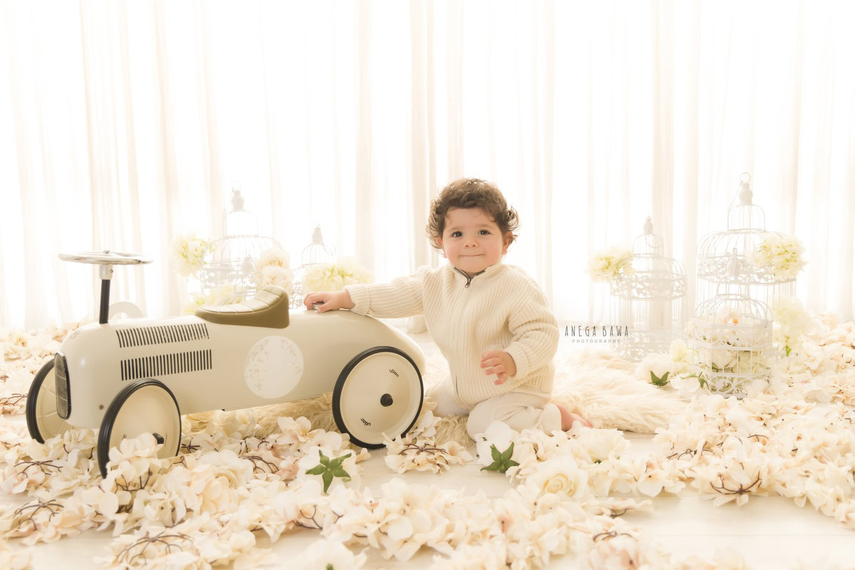 Witness the adorable innocence of a 9-month-old boy, grasping onto his cherished toy scooter amidst a serene backdrop of white flowers and castles. This delightful scene was expertly captured during a toddler photoshoot in Delhi, Gurgaon, and Noida by the talented photographer, Anega Bawa.
