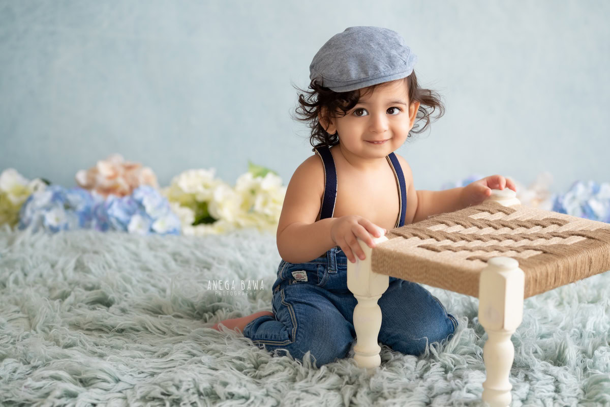 Make memories last with Anega Bawa Photography's sitter photoshoot in Delhi and Gurgaon. Your 9-month-old boy shines in cute cap and dungaree poses, holding a woven stool against a charming blue backdrop and a soft grey rug.