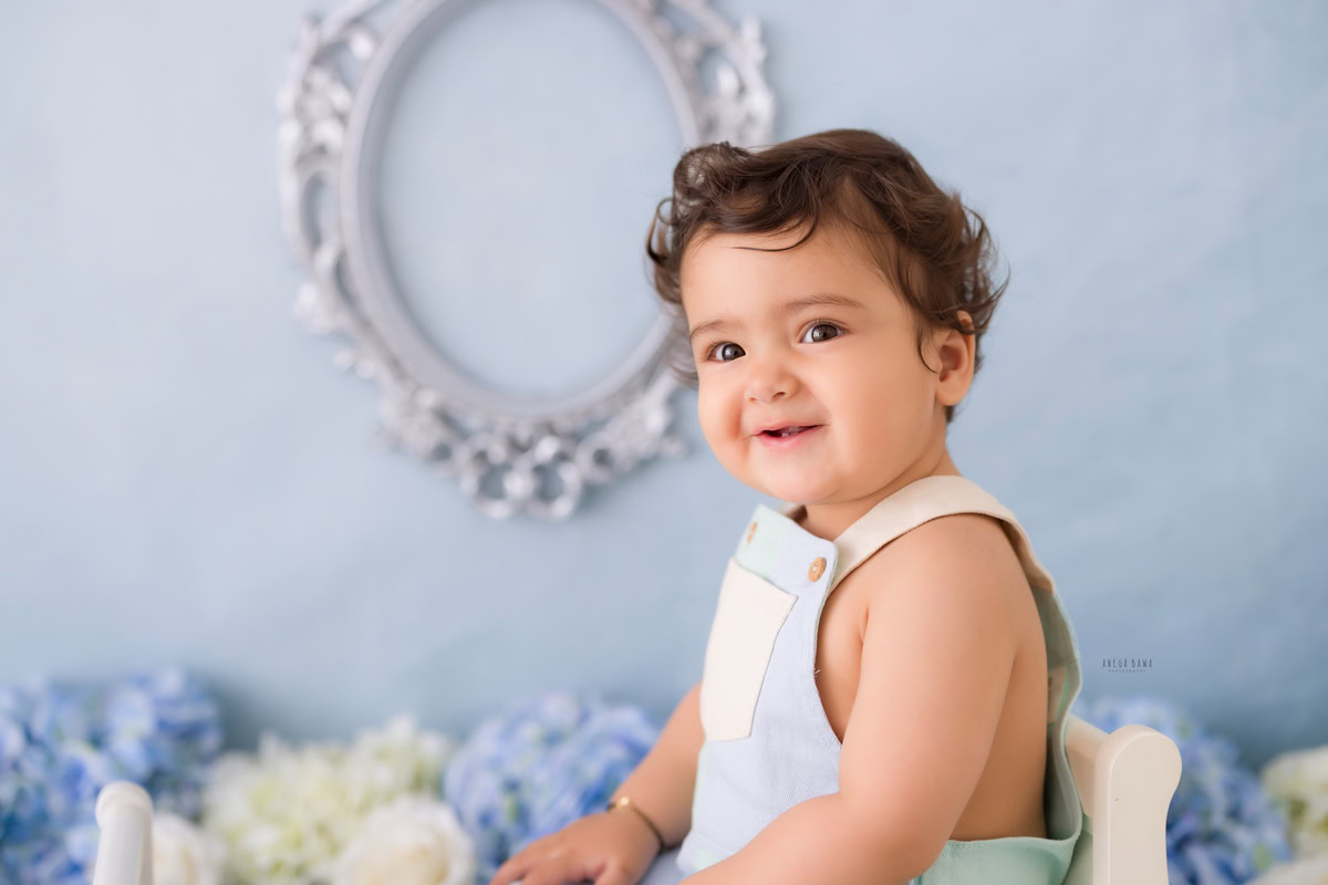 Experience the charm of a 10-month-old boy dressed in blue dungarees, surrounded by blue and white flowers against a striking blue backdrop. This captivating moment was artfully captured during a toddler photoshoot in Delhi, Gurgaon, and Noida by the skilled photographer, Anega Bawa.