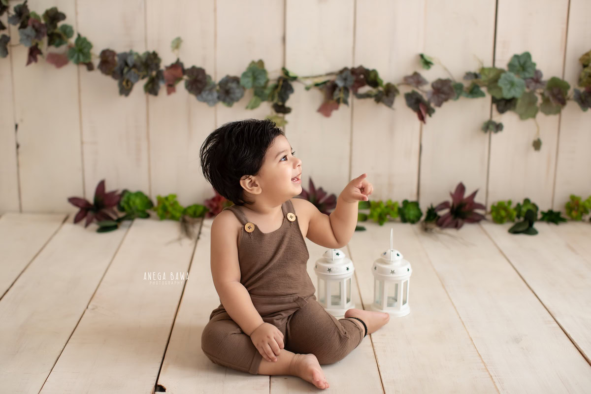 Captured during a delightful sitter photo shoot in Delhi, Gurgaon by Anega Bawa, this charming image features a 10-month-old boy adorned in a brown dungaree. Against a soothing beige backdrop, he sits surrounded by a leafy fringe on the wall, adding a touch of natural beauty to the scene.