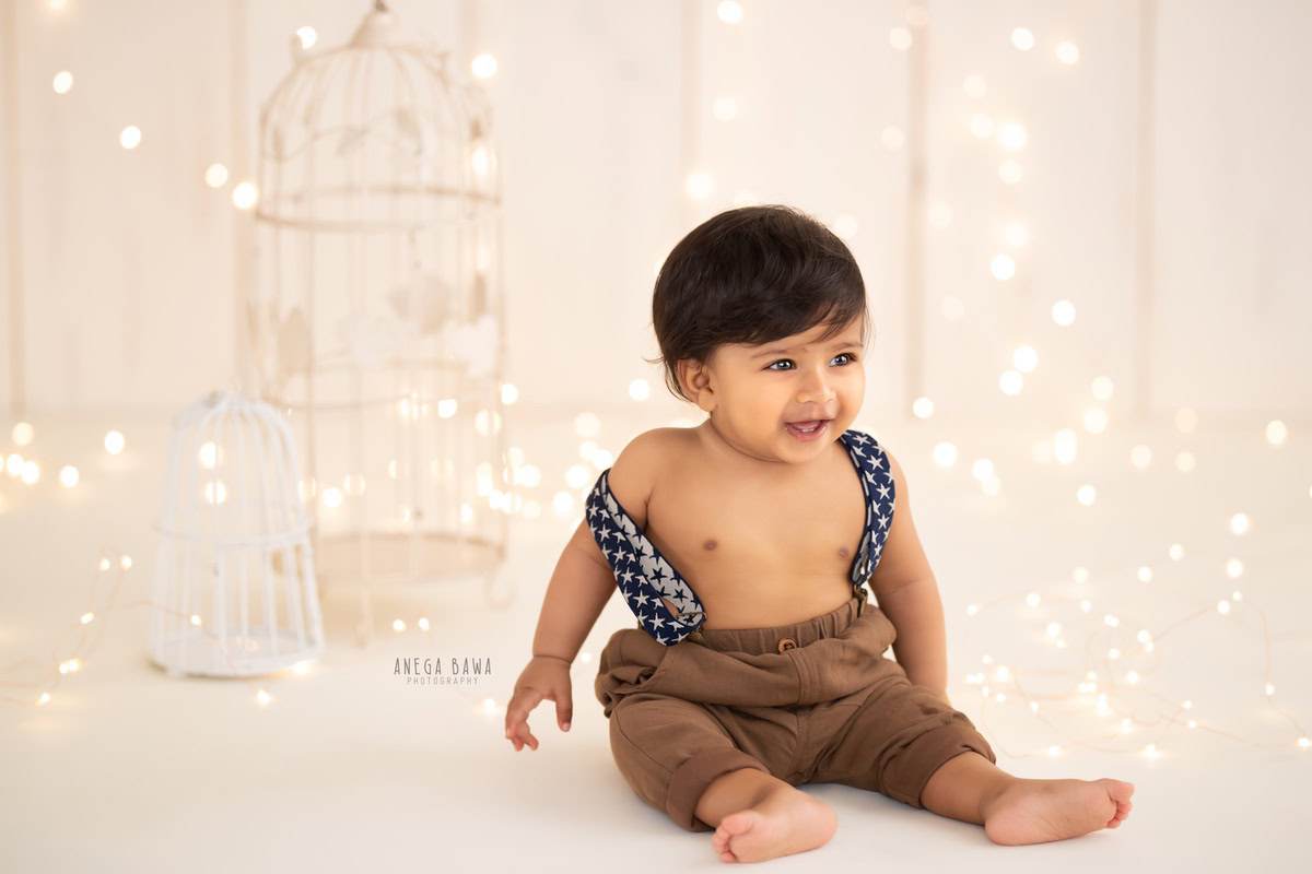 Celebrate your little boy's milestone with Anega Bawa Photography's sitter photoshoot in Delhi and Gurgaon. Adorned in a charming brown dungaree, he sits against a white backdrop embellished with white castles and fairy lights, capturing the innocence and joy of his 10-month-old stage.