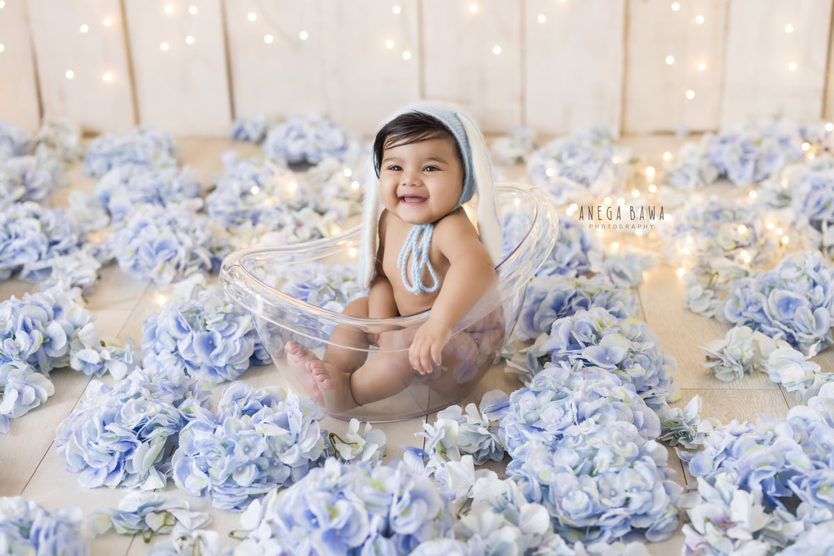 Enchanting boy nestled in a crystal tub surrounded by blue flowers against a serene beige backdrop, bathed in golden lights and adorned with a charming blue cap, capturing the magic of his 10 months first birthday photoshoot in Delhi by Anega Bawa, Gurgaon, Noida