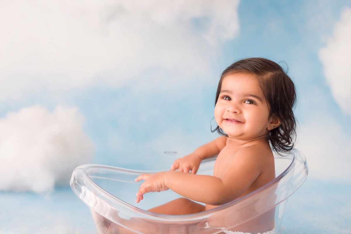 In this adorable snapshot by Anega Bawa from a baby pre-birthday photoshoot in Delhi, a 10-month-old boy beams with joy while seated in a crystal-clear tub against a dreamy cloudy backdrop. His smiling pose radiates pure delight, capturing a precious moment filled with innocence and happiness.