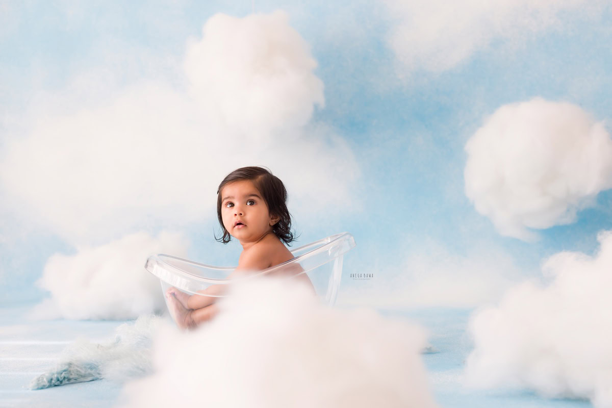 For this captivating shot from a baby pre-birthday photoshoot in Delhi by Anega Bawa, a 10-month-old boy sits contentedly in a crystal-clear tub against a backdrop of fluffy clouds. This enchanting image captures the innocence and wonder of childhood, creating a timeless memory to cherish.
