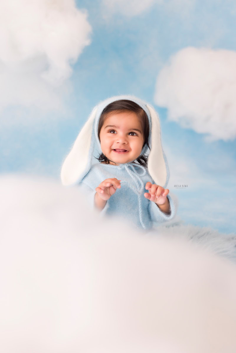 In this delightful sitter birthday photoshoot captured by Anega Bawa in Delhi, a 10-month-old boy steals the spotlight with his adorable blue cap. Against the backdrop of fluffy clouds, his innocent charm shines through, creating a whimsical and enchanting atmosphere. The blue cap adds a pop of color and a touch of playfulness to the scene, perfectly complementing the joyous occasion.
