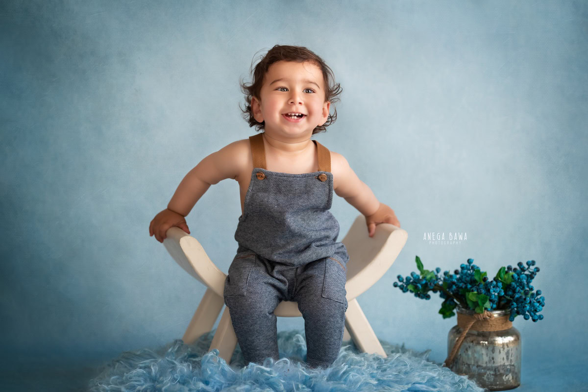 In this adorable capture from a sitter photo shoot in Delhi, Gurgaon by Anega Bawa, a 10-month-old boy dons a trendy denim dungaree. Set against a vibrant blue backdrop, he sits comfortably on a matching blue rug, adding a pop of color to the scene.