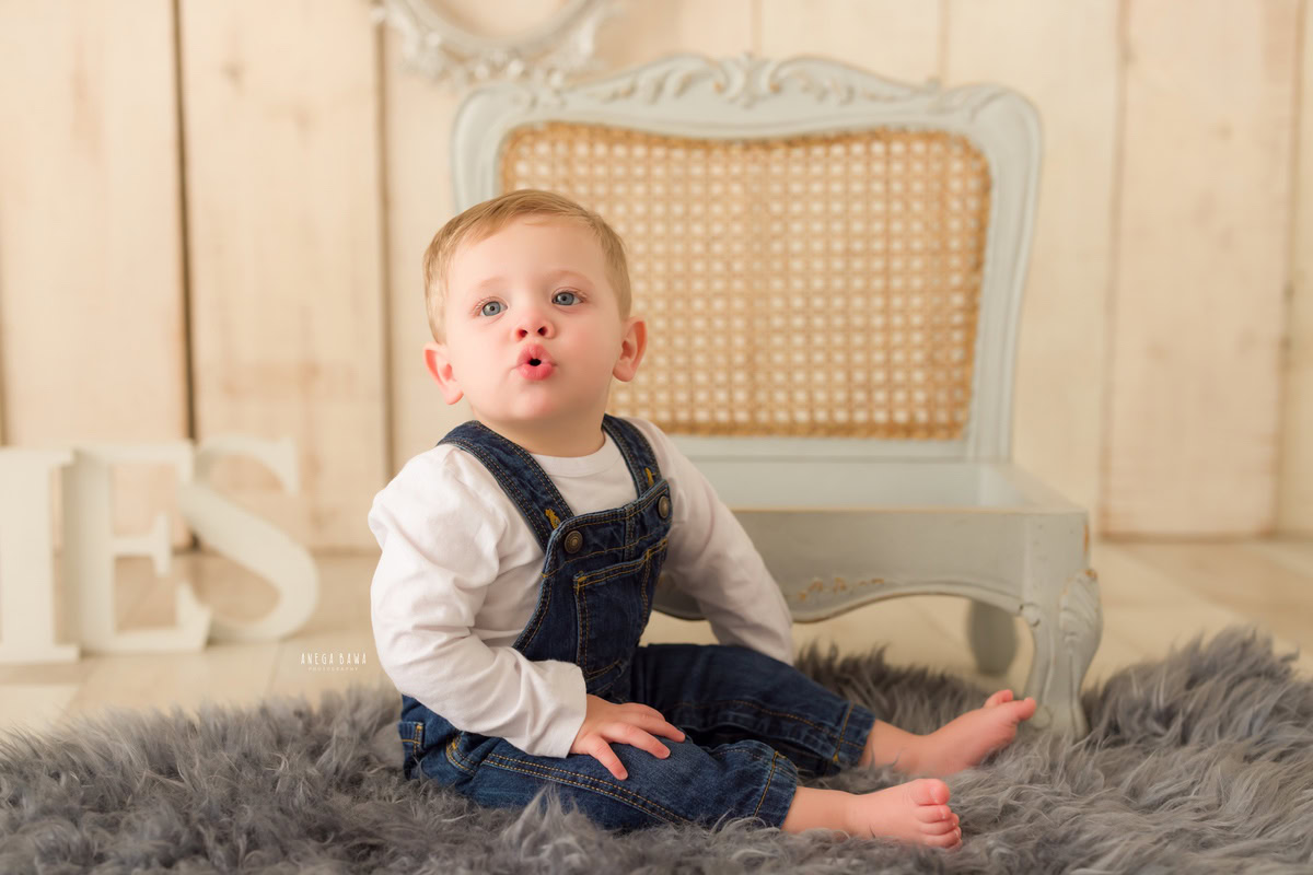 In this captivating baby pre-birthday photoshoot by Anega Bawa in Delhi, a charming 11-month-old boy strikes a playful pout pose while seated on a cozy grey rug and a stylish grey chair. Against a soothing beige backdrop, his adorable expression and relaxed demeanor are beautifully captured, creating timeless memories to cherish.