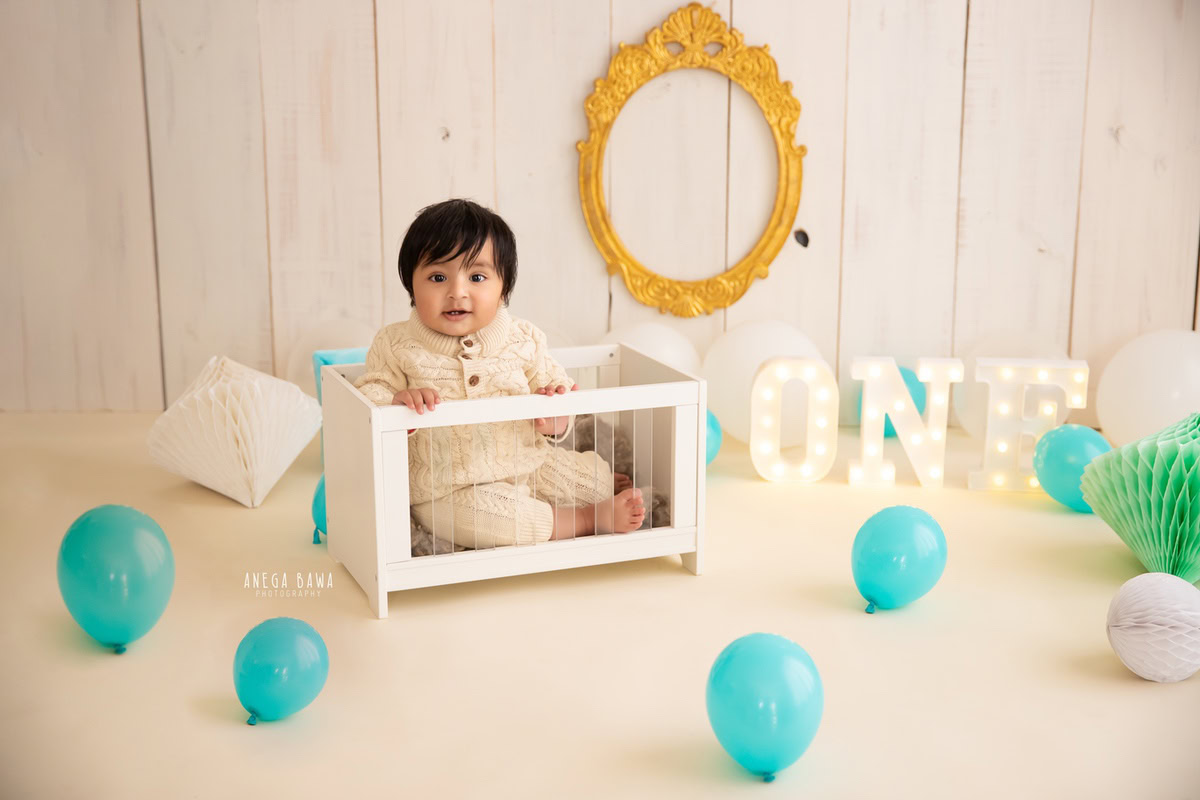 Photographed by sitter photographer Anega Bawa in Delhi, a charming 11-month-old boy sits in a white cot amidst a whimsical scene of blue balloons and a golden frame against a serene beige backdrop.