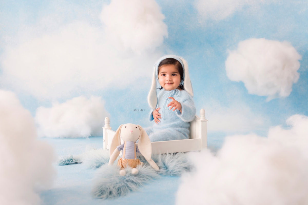 Adorable toddler sitting in a white bed with a plush bunny toy, surrounded by fluffy clouds and a blue sky backdrop, perfect for baby and toddler photography sessions.