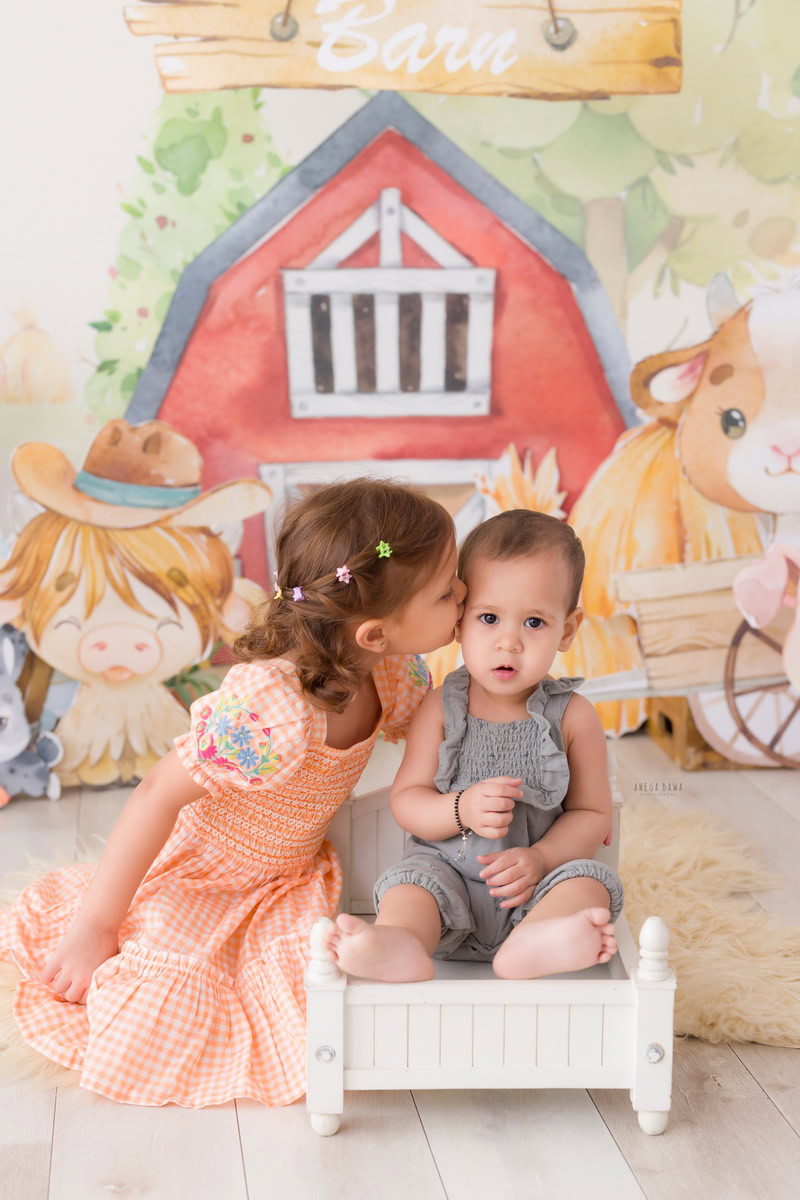 In this heartwarming baby pre-birthday photoshoot by Anega Bawa in Delhi, a 11-month-old boy shares a sweet kissing pose with his sibling while seated in a cozy white cot. The adorable moment is captured against a playful cartoon backdrop, creating a magical atmosphere filled with love and joy.
